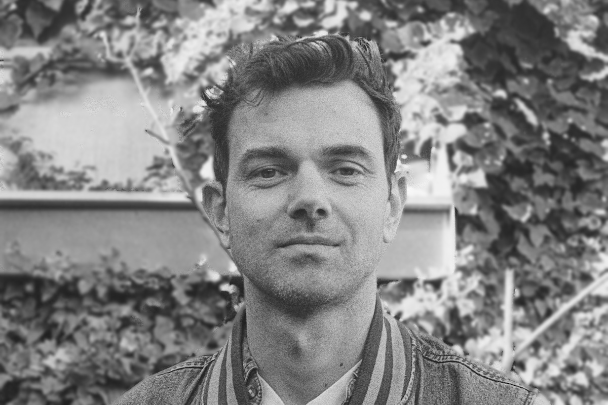 Black-and-white portrait of Tom Iansek from musical project #1 Dads smiling against a leafy garden backdrop.